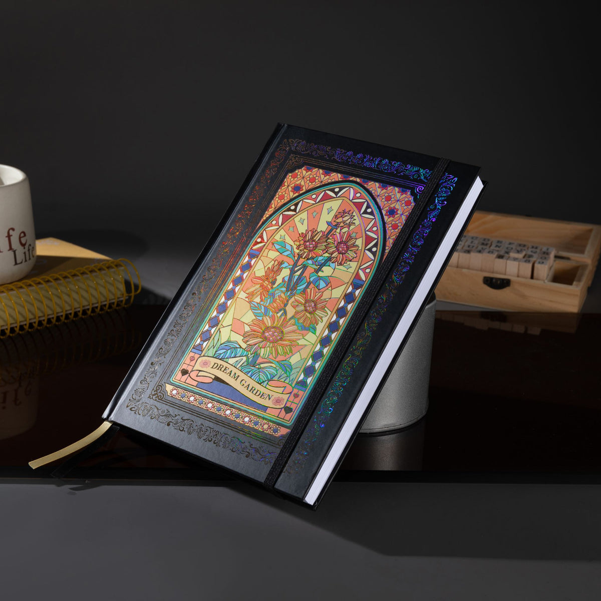Black and gold holographic sunflower Dream Garden cardboard notebook standing angled on a dark reflective surface, featuring a vibrant floral arch design with holographic iridescent borders, styled with a coffee mug, wooden stamps, and a gold ribbon bookmark.