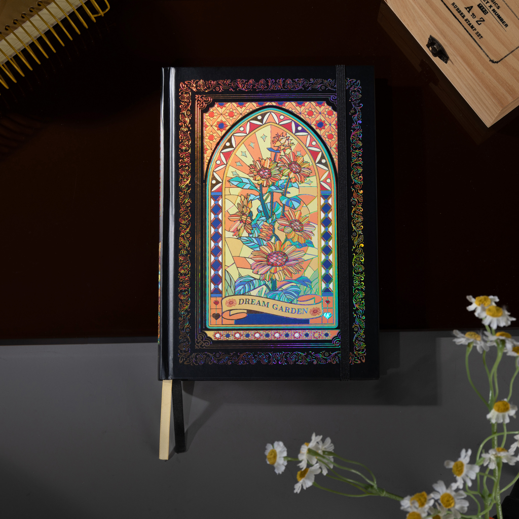Black and gold holographic sunflower Dream Garden cardboard notebook standing upright on a dark surface, featuring a vibrant floral arch design with iridescent borders and a black elastic closure, styled with small white daisy flowers and a wooden stamp box.