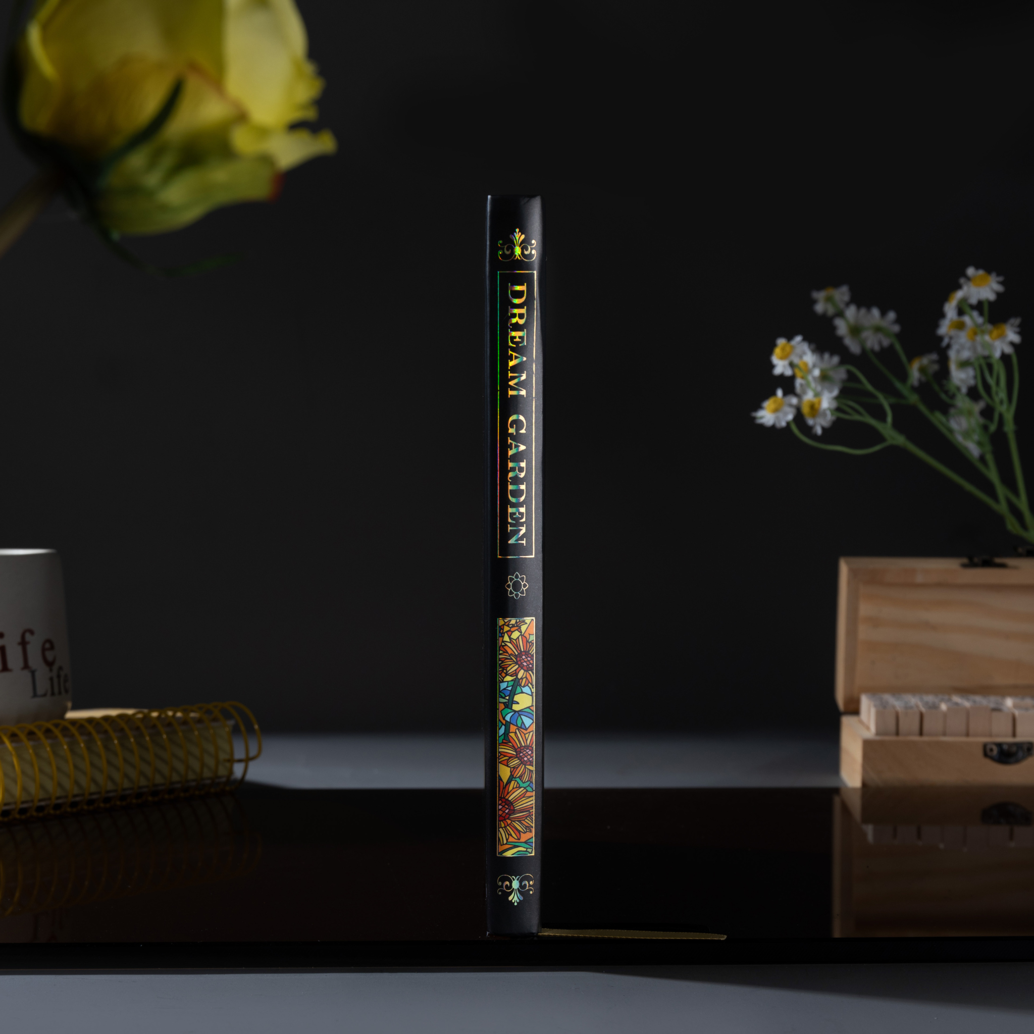 Spine view of the black sunflower Dream Garden cardboard notebook standing upright on a dark surface, featuring the holographic "DREAM GARDEN" title and floral details.
