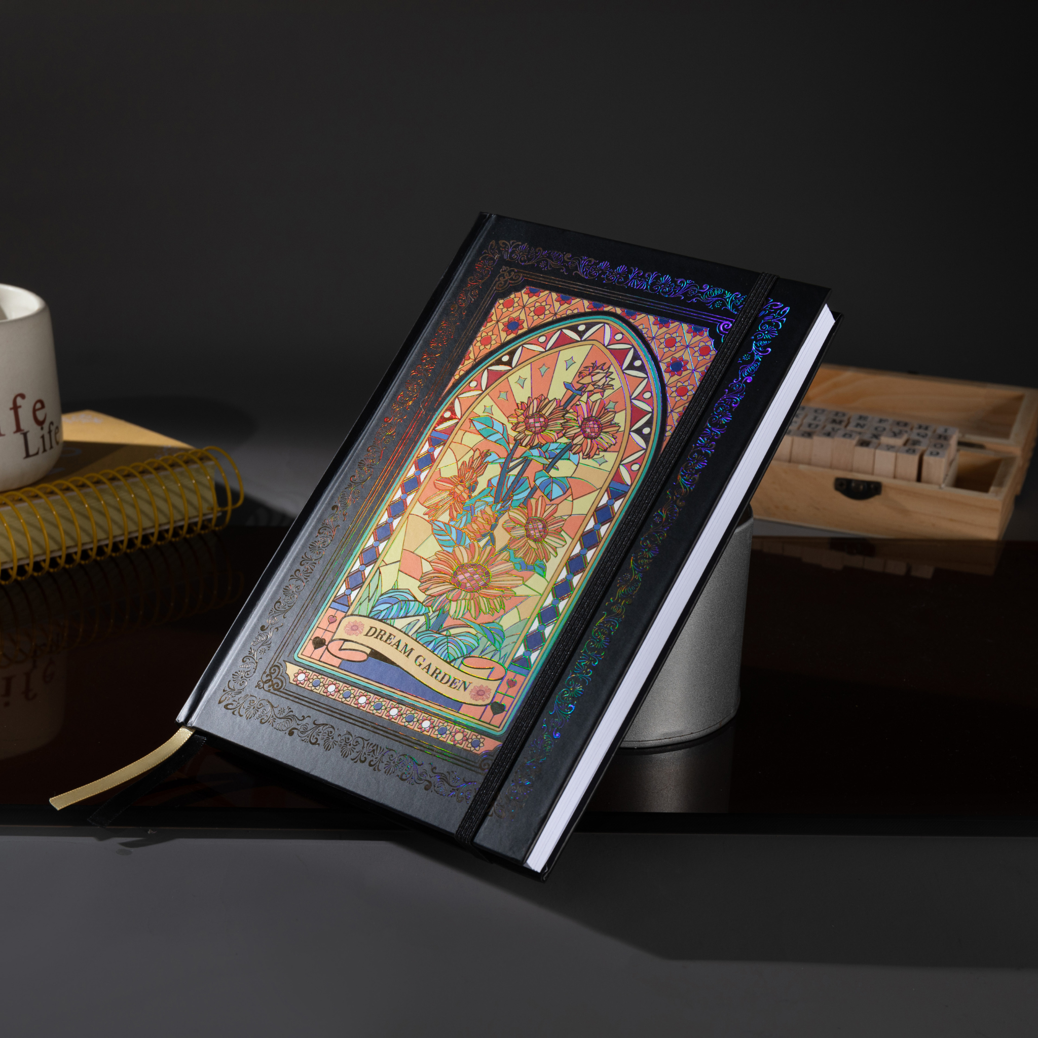 Black and gold holographic sunflower Dream Garden cardboard notebook standing angled on a dark reflective surface, featuring a vibrant floral arch design with holographic iridescent borders, styled with a coffee mug, wooden stamps, and a gold ribbon bookmark.