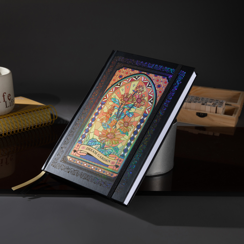 Black and gold holographic sunflower Dream Garden cardboard notebook standing angled on a dark reflective surface, featuring a vibrant floral arch design with holographic iridescent borders, styled with a coffee mug, wooden stamps, and a gold ribbon bookmark.