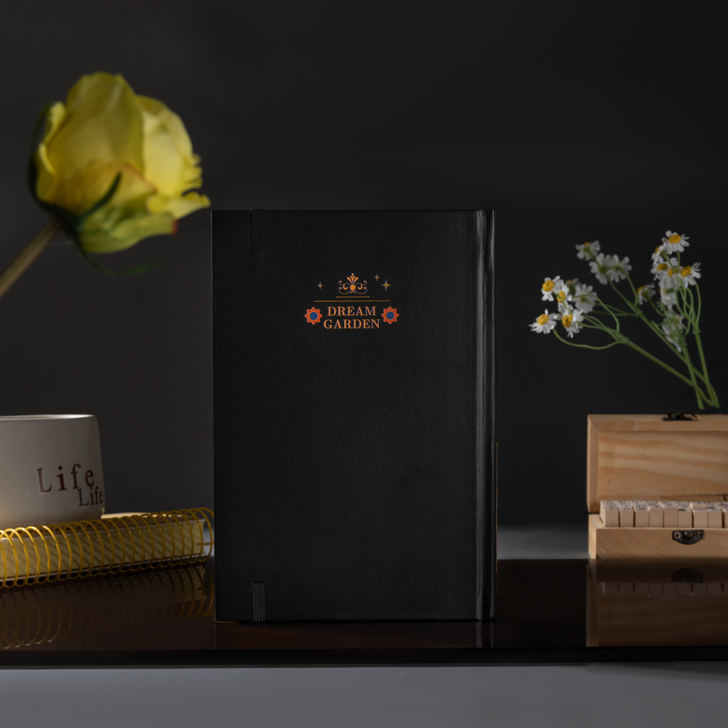 Back cover of the black Dream Garden cardboard notebook standing upright, featuring a minimalist "DREAM GARDEN" logo and star design, styled with dried flowers on a dark surface.