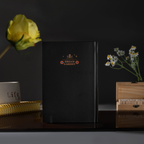 Back cover of the black Dream Garden cardboard notebook standing upright, featuring a minimalist "DREAM GARDEN" logo and star design, styled with dried flowers on a dark surface.