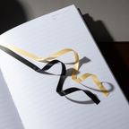 Close-up of the black and gold two-tone ribbon bookmark resting elegantly on the open lined pages of the sunflower Dream Garden notebook.