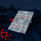Angled flat lay of the blue floral woven fabric notebook resting on a dark blue shimmering fabric surface, featuring an intricate botanical pattern, a matching blue elastic band, and styled with bright red winter berries.