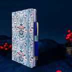 Side view of the blue floral fabric notebook standing upright, showing the side pen loop holding a blue and gold pen, styled on dark fabric with red berries.