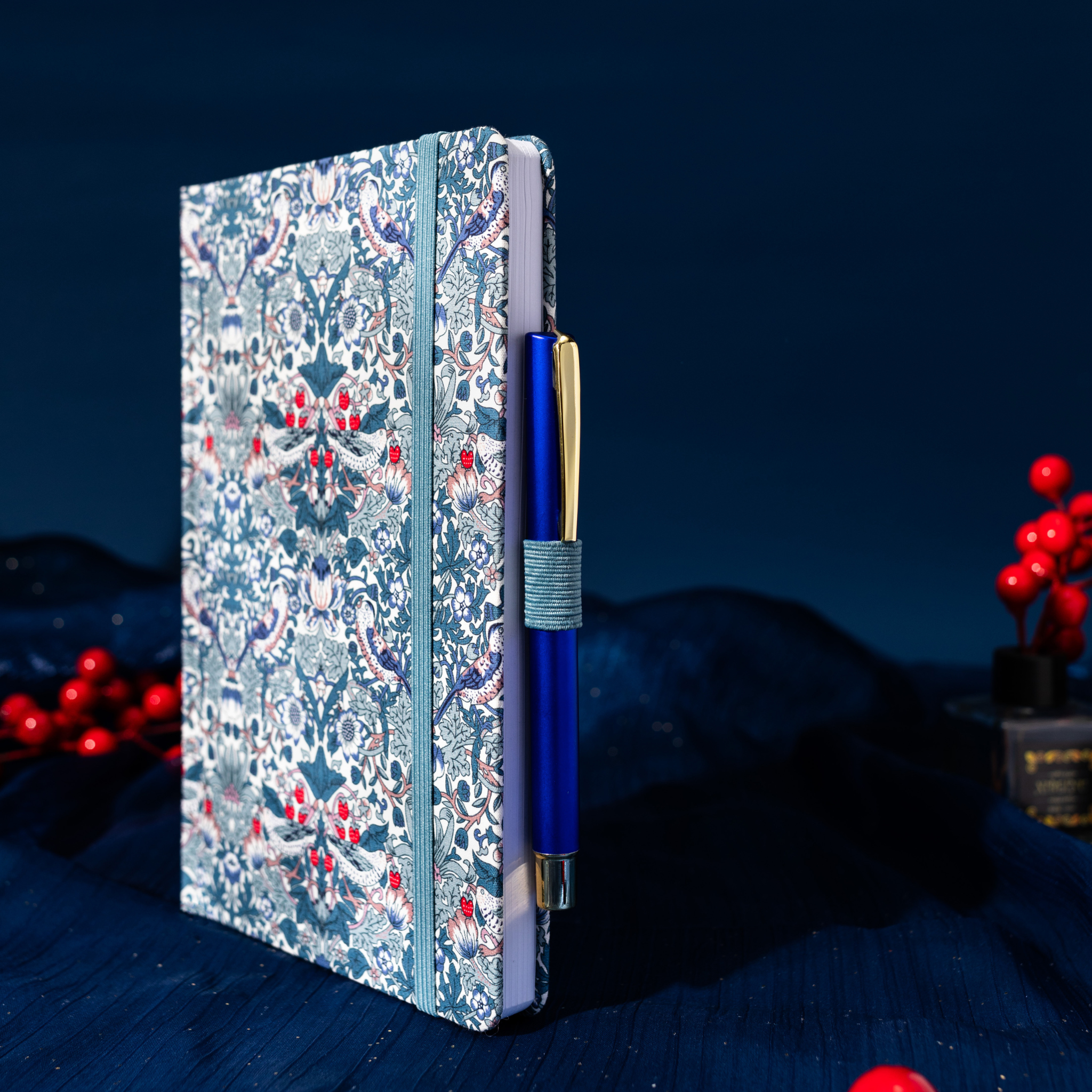 Side view of the blue floral fabric notebook standing upright, showing the side pen loop holding a blue and gold pen, styled on dark fabric with red berries.