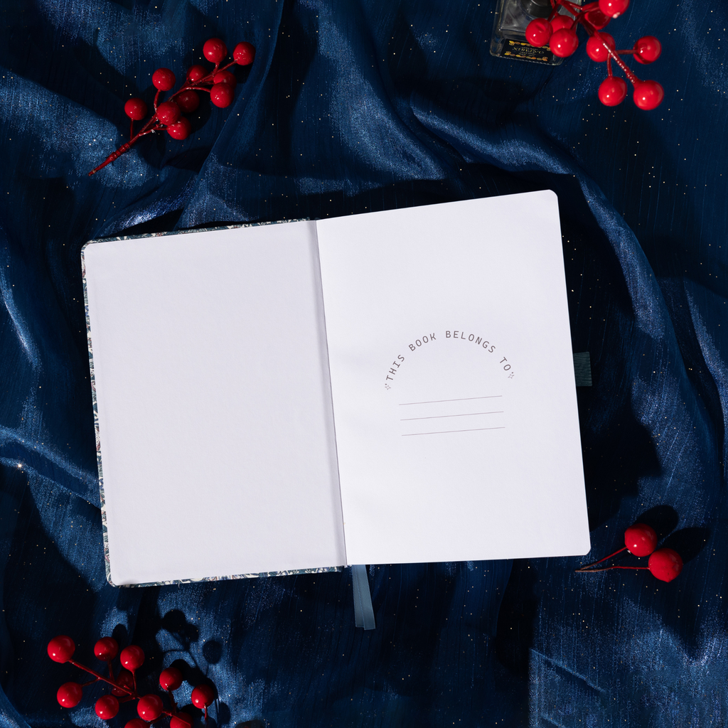 Open blue floral notebook showing the "THIS BOOK BELONGS TO" ownership page with decorative text and lines, styled on dark blue fabric with red berries.