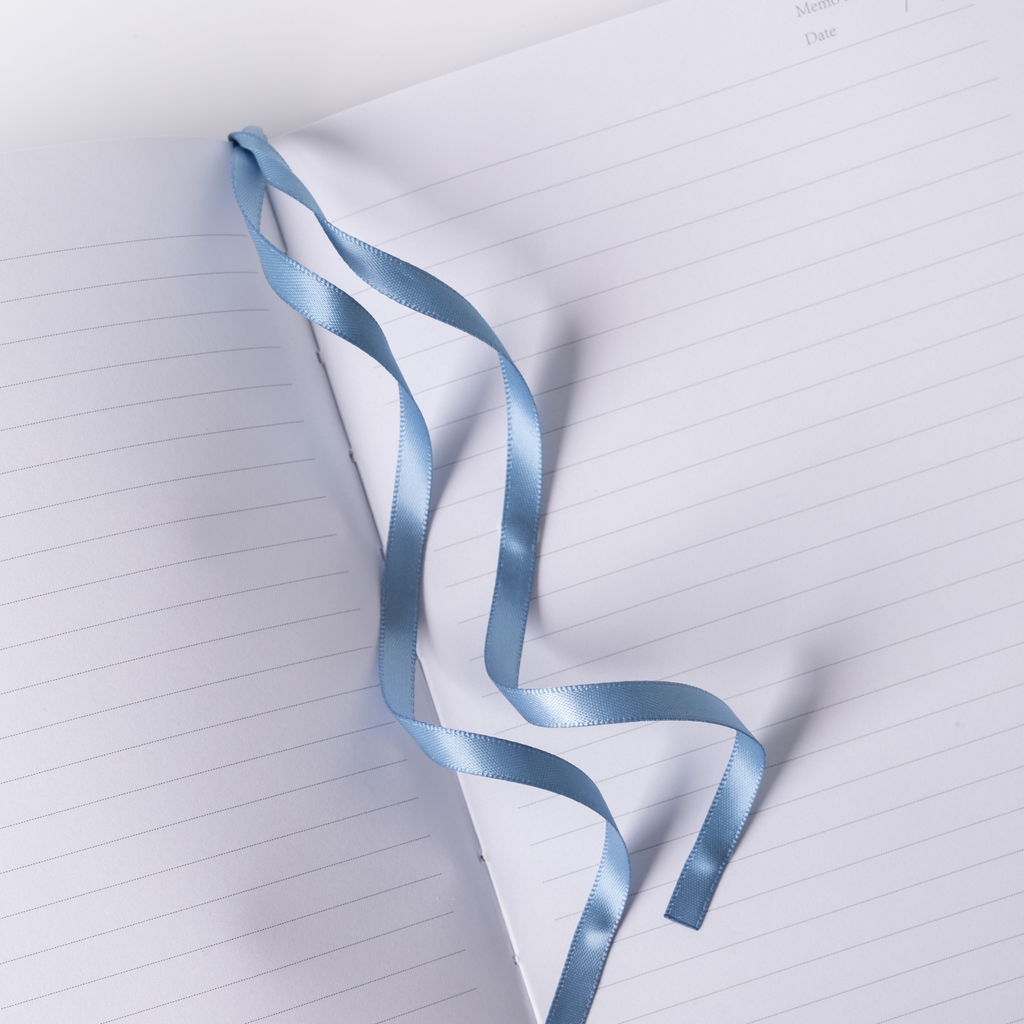 Light blue satin ribbon bookmark resting on the open lined pages of the blue floral notebook, styled on dark blue fabric.