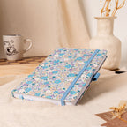 A5 lilac and blue ditsy floral fabric notebook angled on a cream linen cloth, featuring a matching blue elastic closure, styled with a coffee mug and a vase of dried flowers in a warm natural light setting.