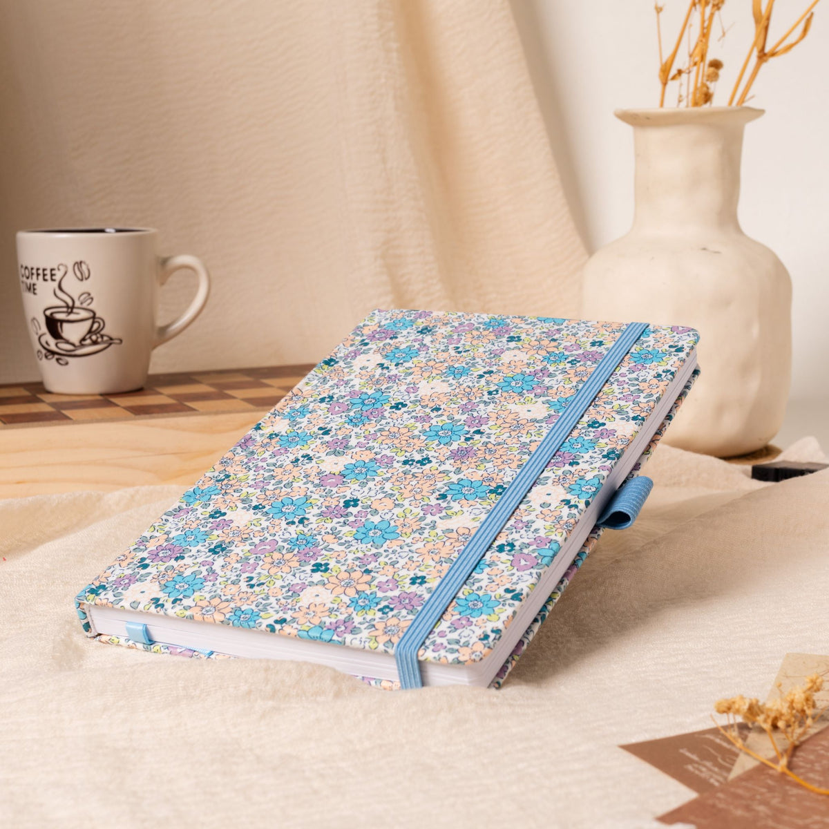 A5 lilac and blue ditsy floral fabric notebook angled on a cream linen cloth, featuring a matching blue elastic closure, styled with a coffee mug and a vase of dried flowers in a warm natural light setting.