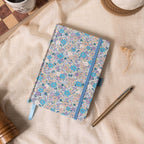 Flat lay of the lilac and blue ditsy floral fabric notebook on a soft cream linen cloth, showcasing the woven floral cover, a matching blue elastic band, a blue ribbon bookmark, and a gold pen, styled with dried flowers.