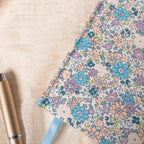 Close-up detail of the lilac and blue ditsy floral fabric notebook cover, showcasing the woven fabric texture, delicate pastel floral pattern, and a light blue ribbon, styled with a gold pen on a soft cream surface.