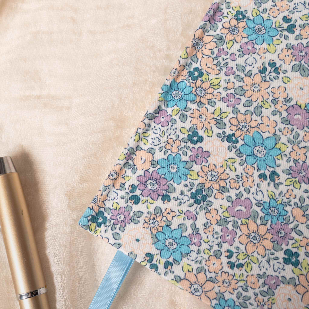 Close-up detail of the lilac and blue ditsy floral fabric notebook cover, showcasing the woven fabric texture, delicate pastel floral pattern, and a light blue ribbon, styled with a gold pen on a soft cream surface.