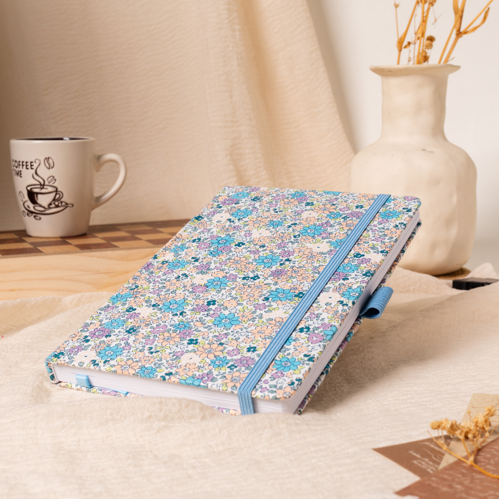 A5 lilac and blue ditsy floral fabric notebook angled on a cream linen cloth, featuring a matching blue elastic closure, styled with a coffee mug and a vase of dried flowers in a warm natural light setting.