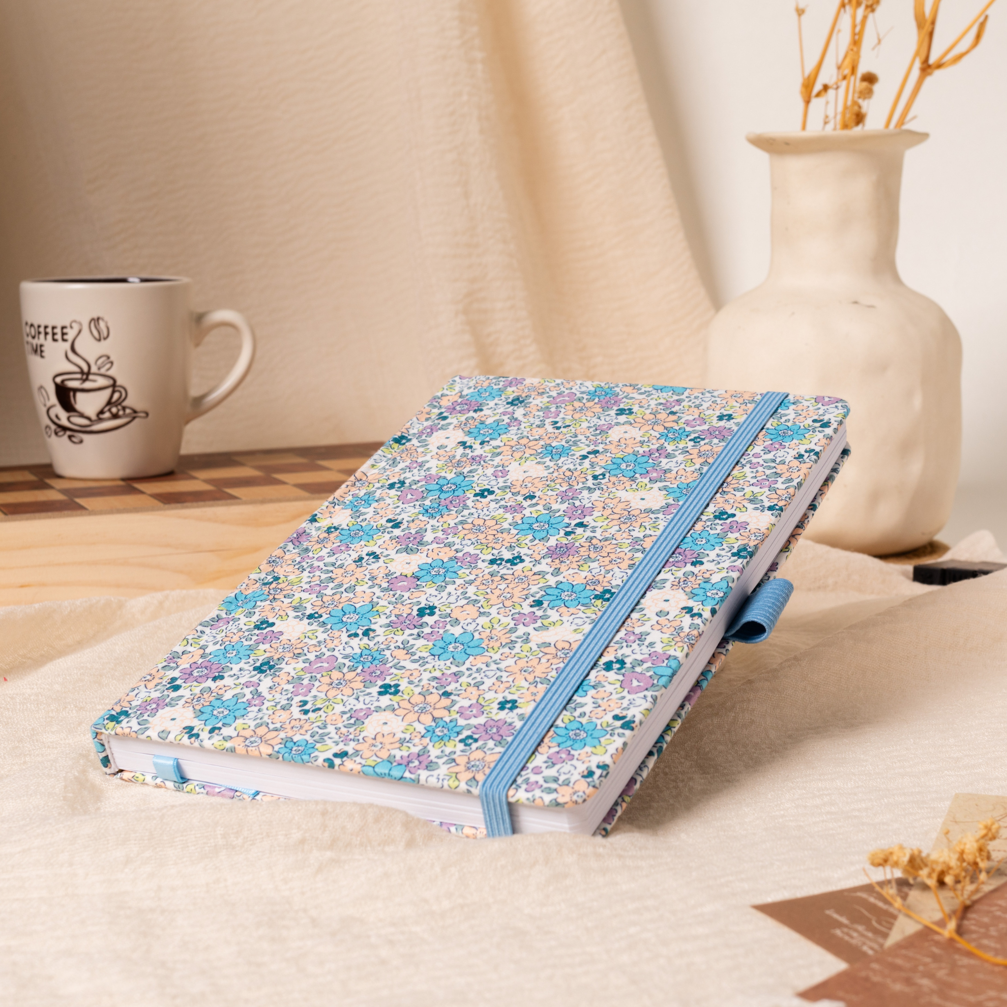 A5 lilac and blue ditsy floral fabric notebook angled on a cream linen cloth, featuring a matching blue elastic closure, styled with a coffee mug and a vase of dried flowers in a warm natural light setting.
