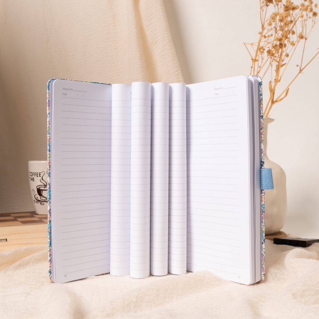 Open lilac and blue floral fabric notebook standing vertically, displaying multiple layered lined pages, a lay-flat binding, and Memo No. and Date headers, styled with a coffee mug and dried flowers on a cream fabric surface.