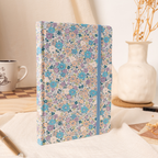 Lilac and blue ditsy floral fabric notebook standing upright on a cream linen surface, styled with dried flowers and a ceramic vase.