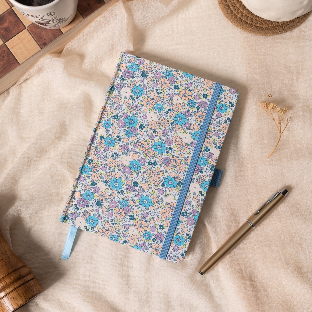 Flat lay of the lilac and blue ditsy floral fabric notebook on a soft cream linen cloth, showcasing the woven floral cover, a matching blue elastic band, a blue ribbon bookmark, and a gold pen, styled with dried flowers.
