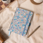 Flat lay of the lilac and blue ditsy floral fabric notebook on a soft cream linen cloth, showcasing the woven floral cover, a matching blue elastic band, a blue ribbon bookmark, and a gold pen, styled with dried flowers.
