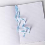 Light blue satin ribbon bookmark elegantly draped across the open lined pages of the lilac and blue ditsy floral notebook.