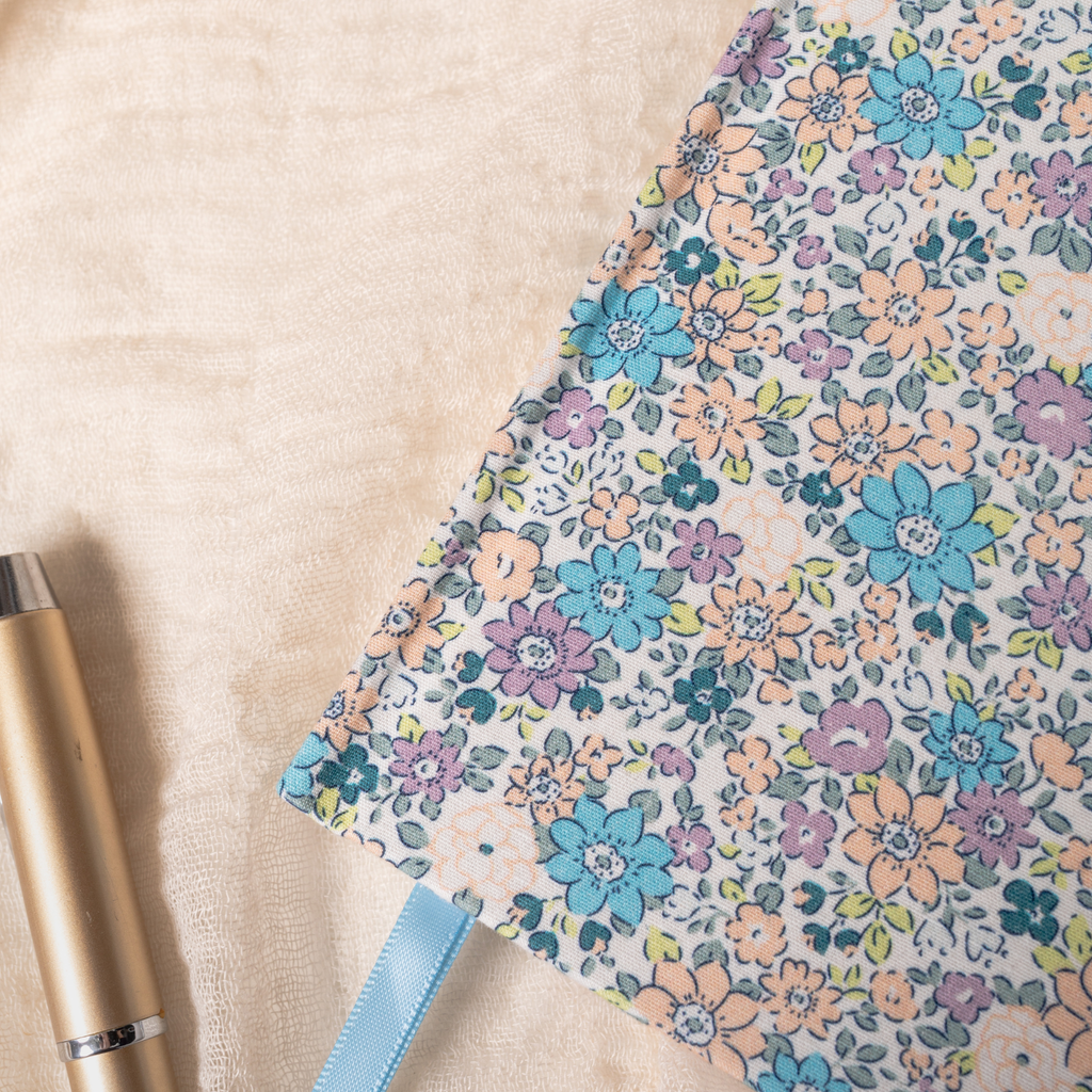 Close-up detail of the lilac and blue ditsy floral fabric notebook cover, showcasing the woven fabric texture, delicate pastel floral pattern, and a light blue ribbon, styled with a gold pen on a soft cream surface.