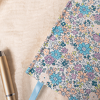 Close-up detail of the lilac and blue ditsy floral fabric notebook cover, showcasing the woven fabric texture, delicate pastel floral pattern, and a light blue ribbon, styled with a gold pen on a soft cream surface.