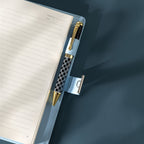 Close-up of the open light blue vegan leather journal, showing a black and white patterned pen tucked into the side pen loop, with the metal lock strap extended, and lined paper with a page number visible.