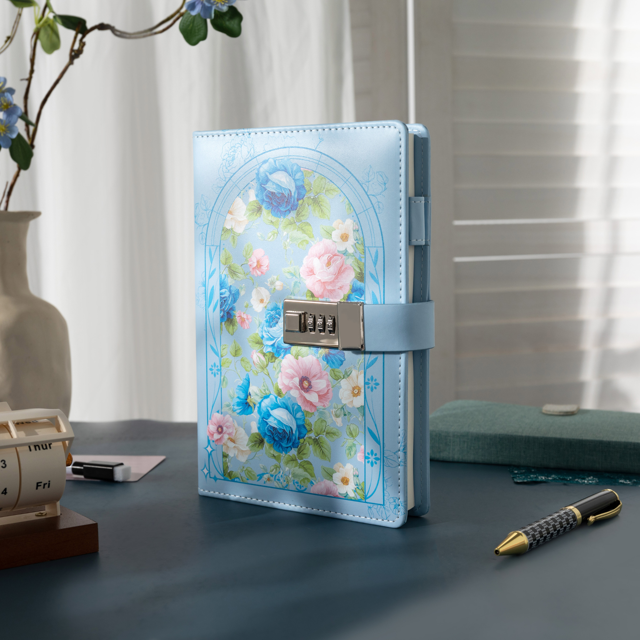 A5 light blue vegan leather notebook with a metal combination lock, featuring a floral arch design with blue and pink roses, standing upright on a dark desk with a wooden calendar and a patterned pen.