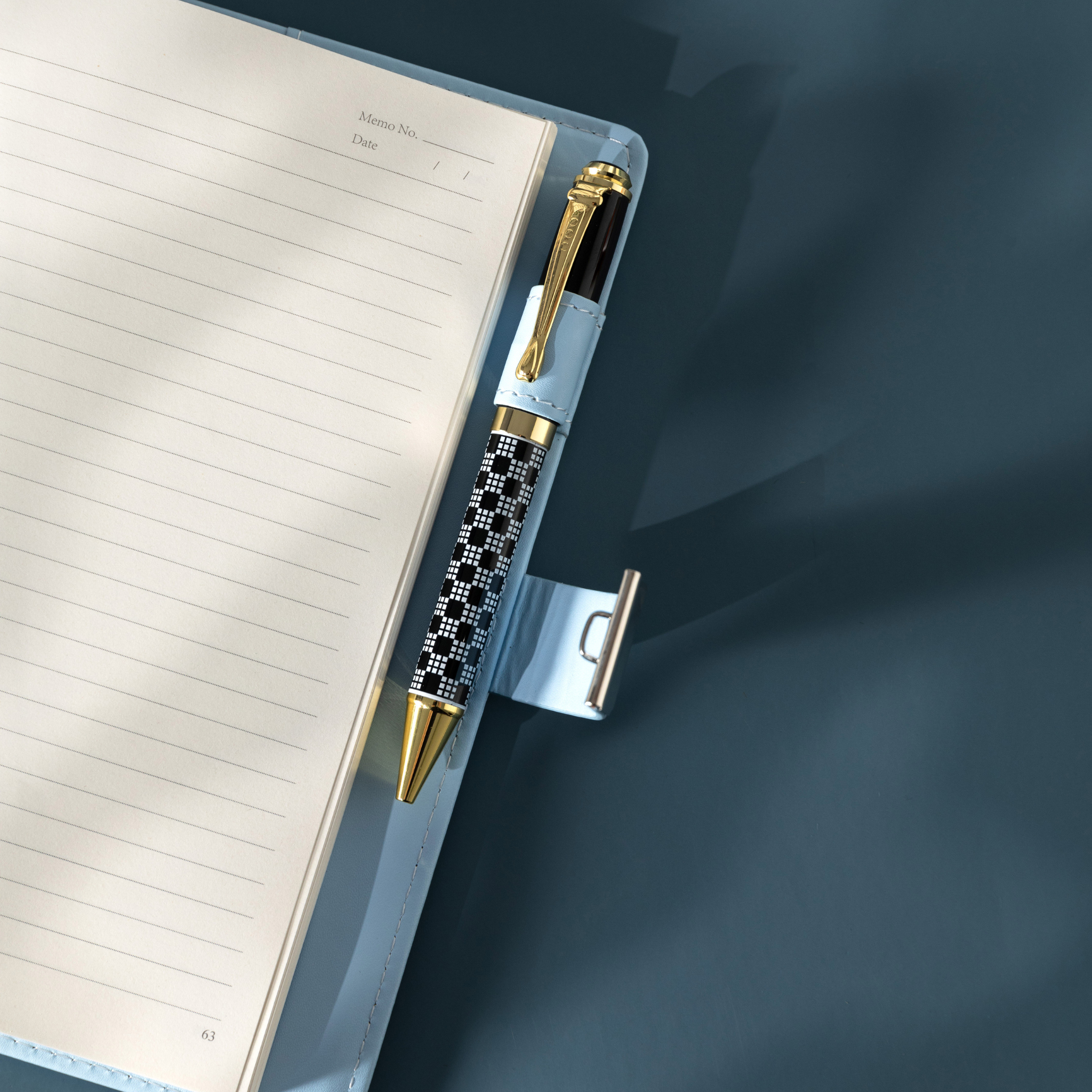 Close-up of the open light blue vegan leather notebook, showing a black and white patterned pen tucked into the side pen loop, with the metal lock strap extended, and lined paper with a page number visible.