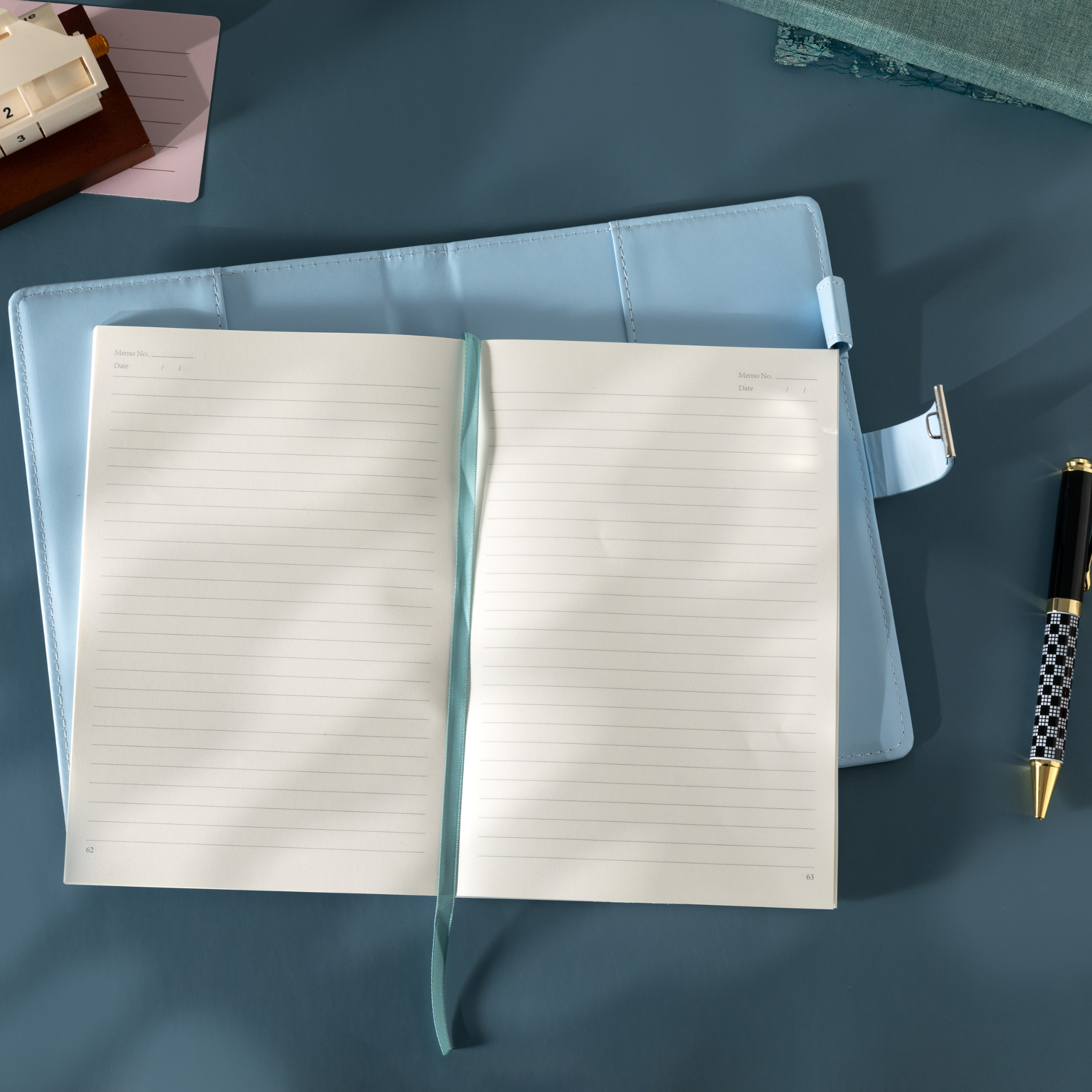 Top-down view of the open light blue floral notebook laid flat, showing the lined paper interior, blue ribbon, and lock strap.