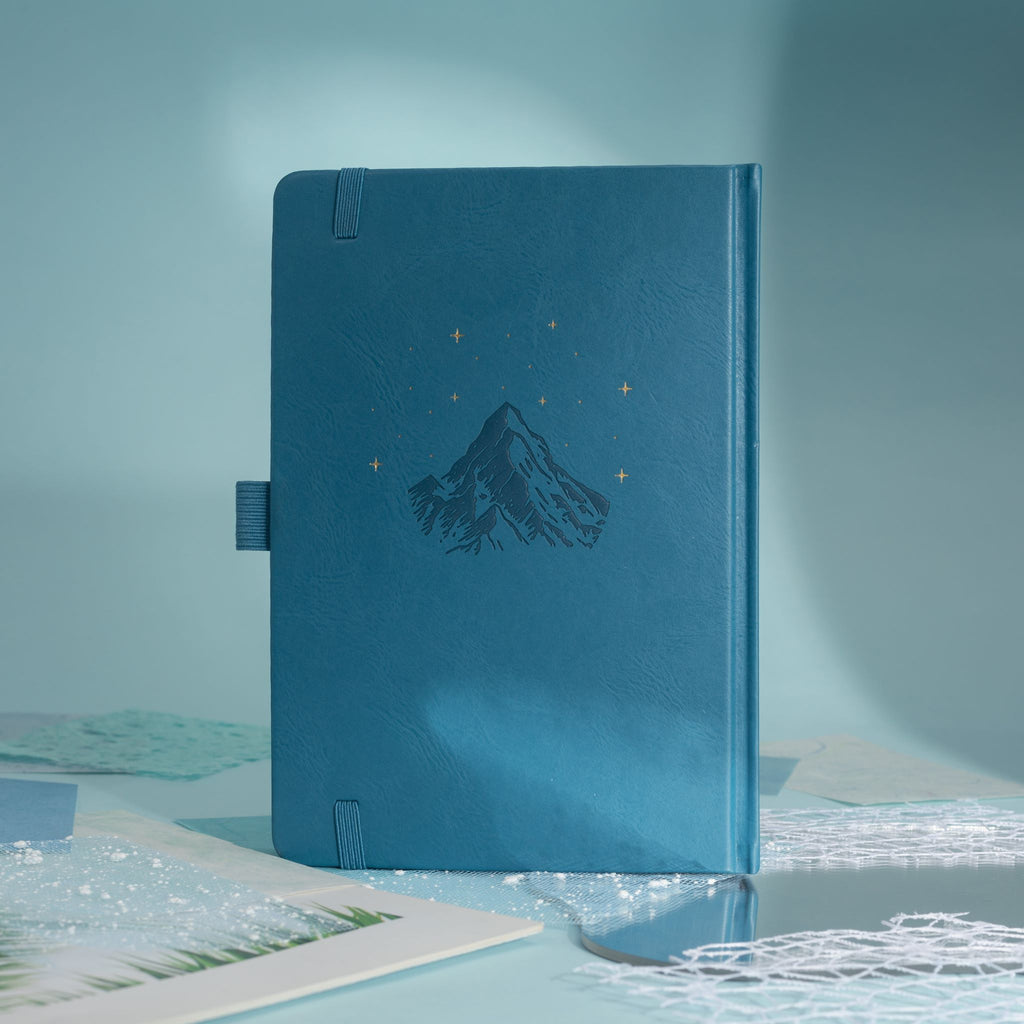 Back cover of the deep blue vegan leather notebook, showcasing a subtle embossed mountain design with small gold foil stars, side pen loop, and matching blue elastic band.