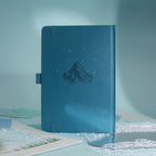 Back cover of the deep blue vegan leather notebook, showcasing a subtle embossed mountain design with small gold foil stars, side pen loop, and matching blue elastic band.