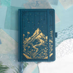 Deep blue vegan leather notebook lying flat on a light wood desk, showcasing a striking gold foil mountain and starry night cover.
