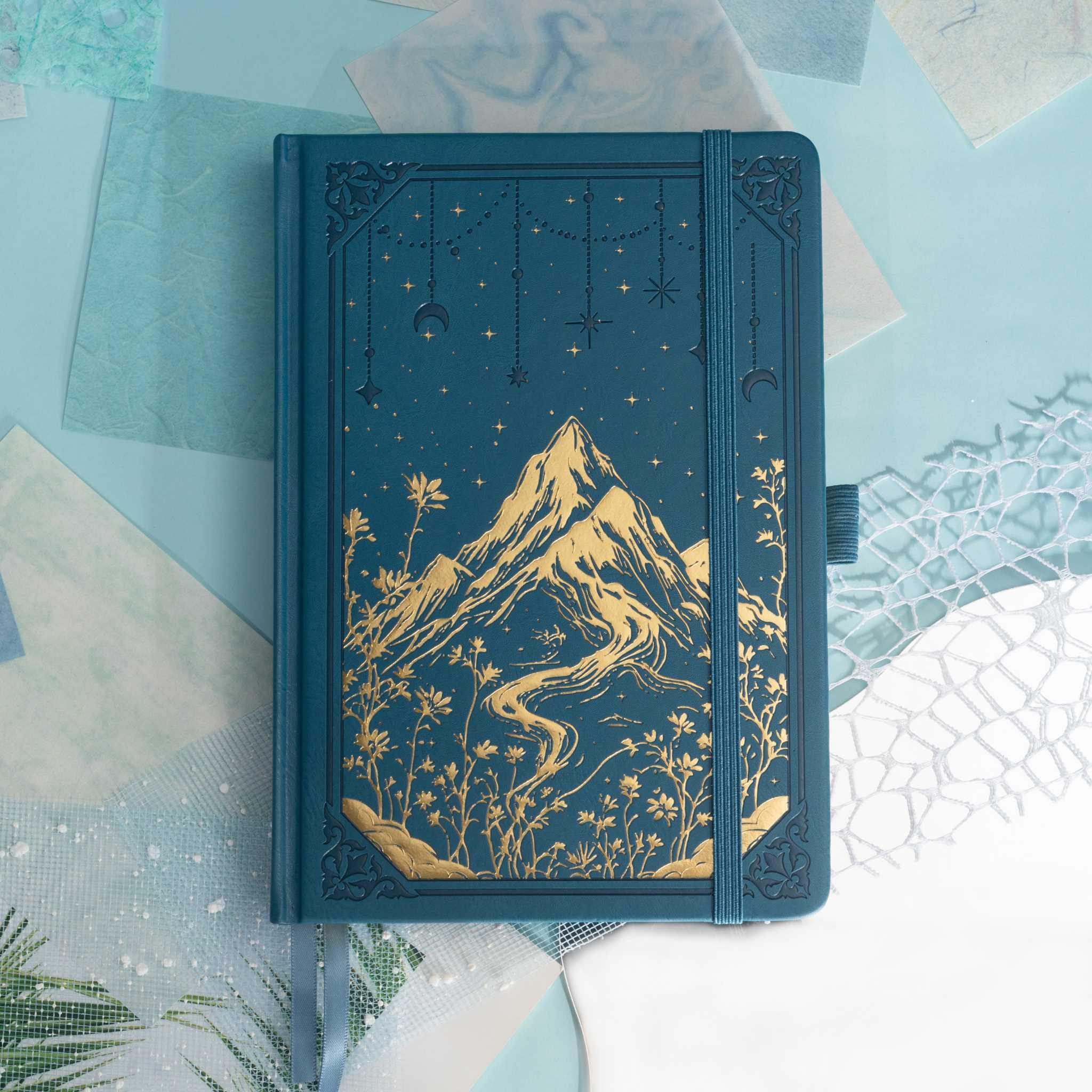 Deep blue vegan leather notebook lying flat on a light wood desk, showcasing a striking gold foil mountain and starry night cover.