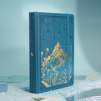 Angled side view of the deep blue vegan leather notebook showcasing the gold foil mountain cover, gold gilded edges, and matching blue elastic band, styled on a light blue surface.