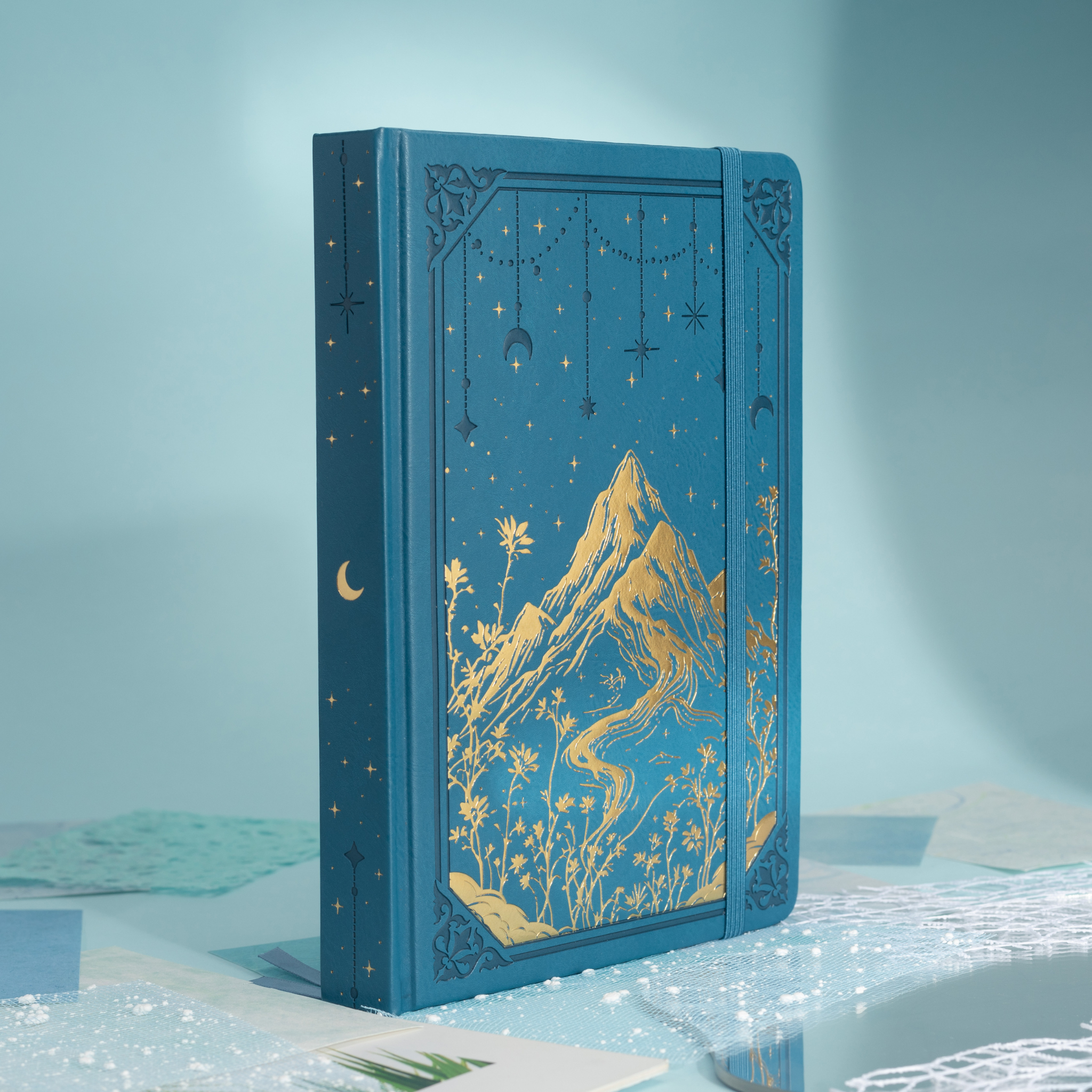 Angled side view of the deep blue vegan leather notebook showcasing the gold foil mountain cover, gold gilded edges, and matching blue elastic band, styled on a light blue surface.