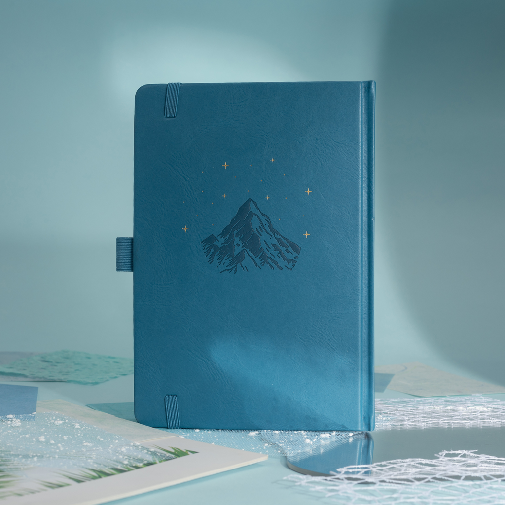 Back cover of the deep blue vegan leather notebook, showcasing a subtle embossed mountain design with small gold foil stars, side pen loop, and matching blue elastic band.