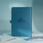 Back cover of the deep blue vegan leather notebook, showcasing a subtle embossed mountain design with small gold foil stars, side pen loop, and matching blue elastic band.