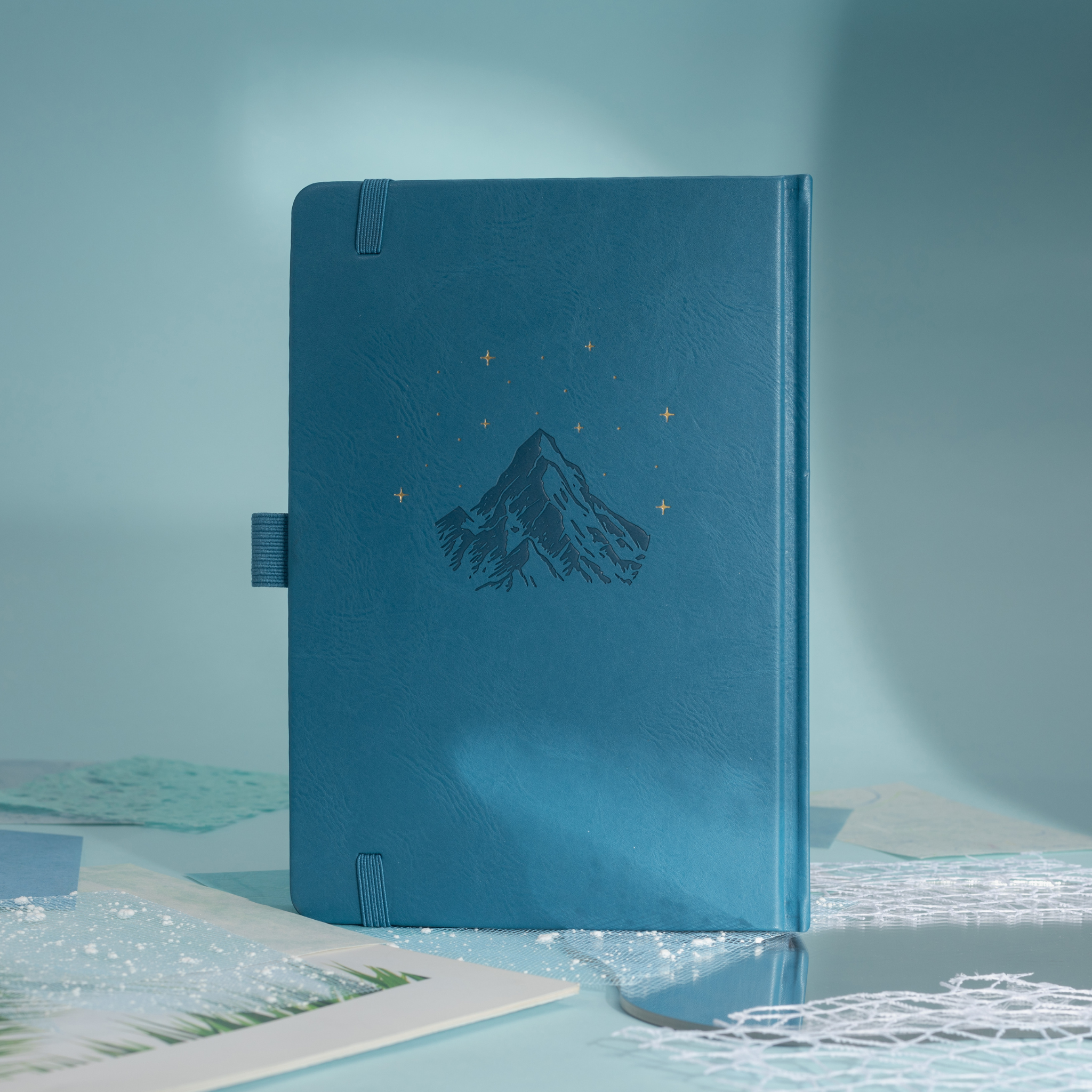 Back cover of the deep blue vegan leather notebook, showcasing a subtle embossed mountain design with small gold foil stars, side pen loop, and matching blue elastic band.