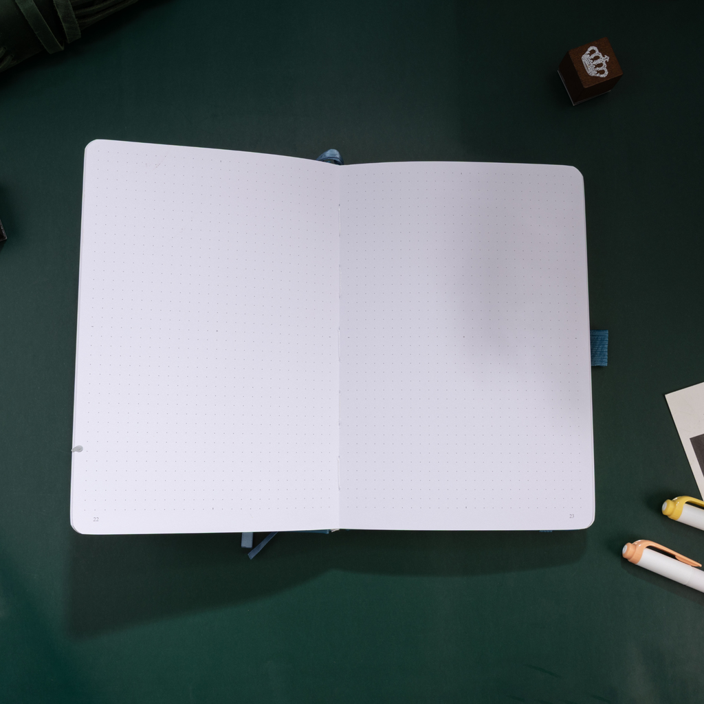 Open A5 journal laid flat on a dark desk, displaying the clean 160gsm dot grid paper interior and a blue ribbon bookmark.