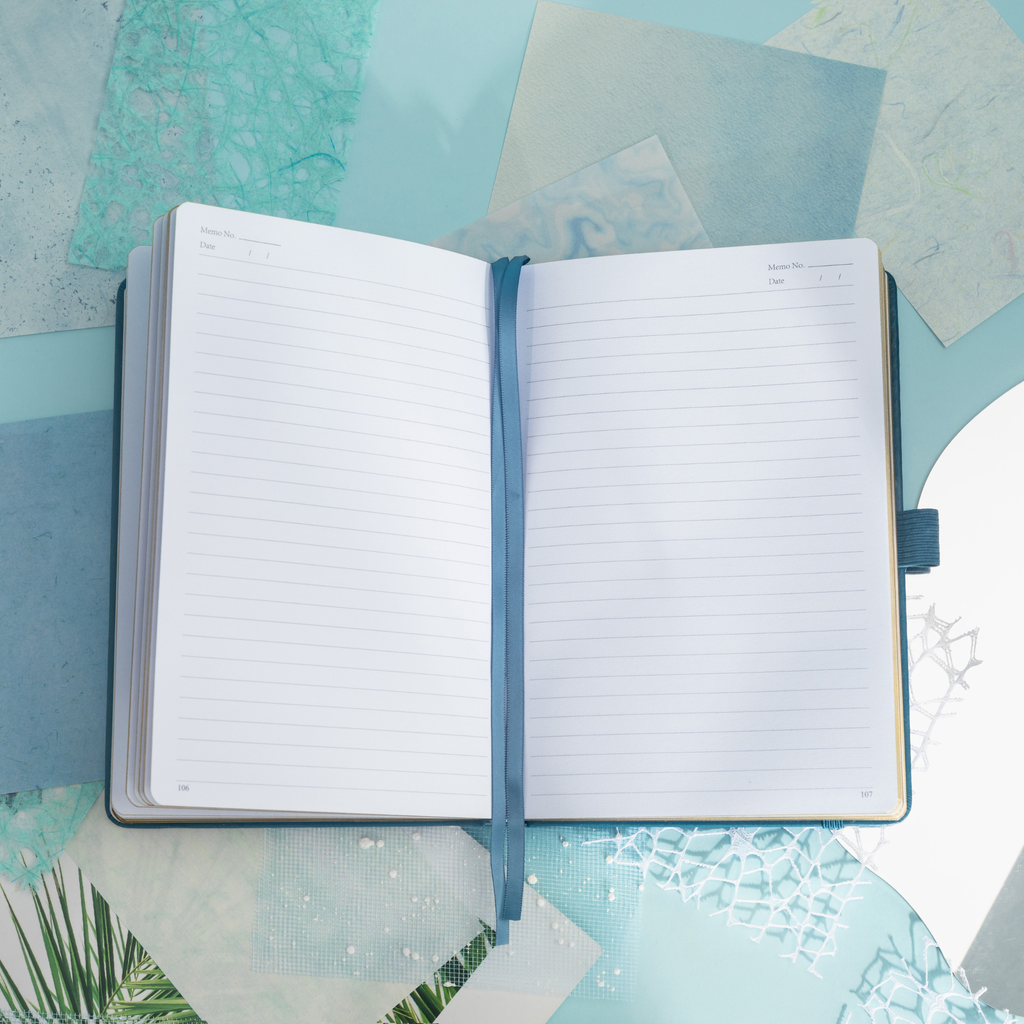 Close-up of the open A5 notebook showing the 100gsm lined paper interior with golden borders, corner numbers, and a blue ribbon bookmark.
