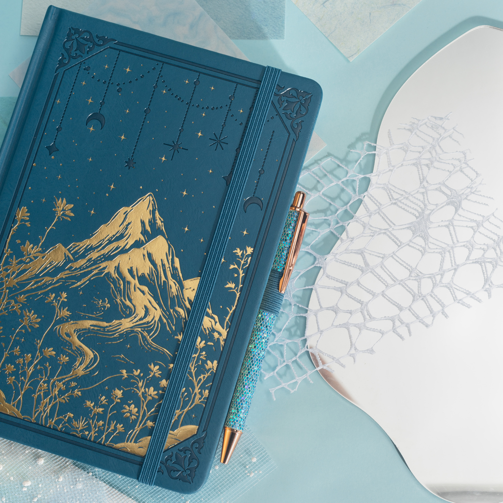 Close-up of the deep blue vegan leather notebook showing the gold foil mountain and starry night cover, paired with a matching blue glitter pen tucked into the side pen loop.