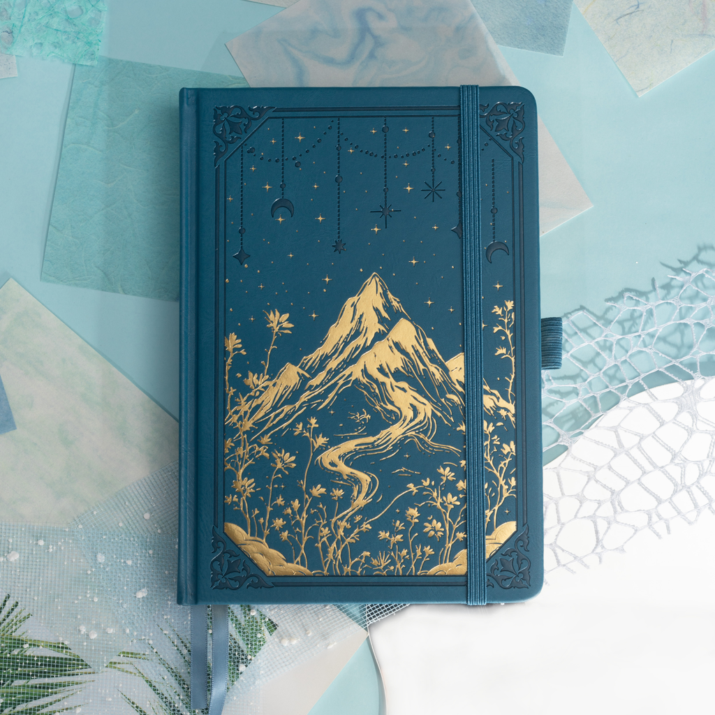 Deep blue vegan leather notebook lying flat on a light wood desk, showcasing a striking gold foil mountain and starry night cover.