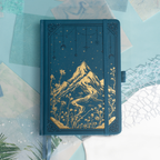 Deep blue vegan leather notebook lying flat on a light wood desk, showcasing a striking gold foil mountain and starry night cover.