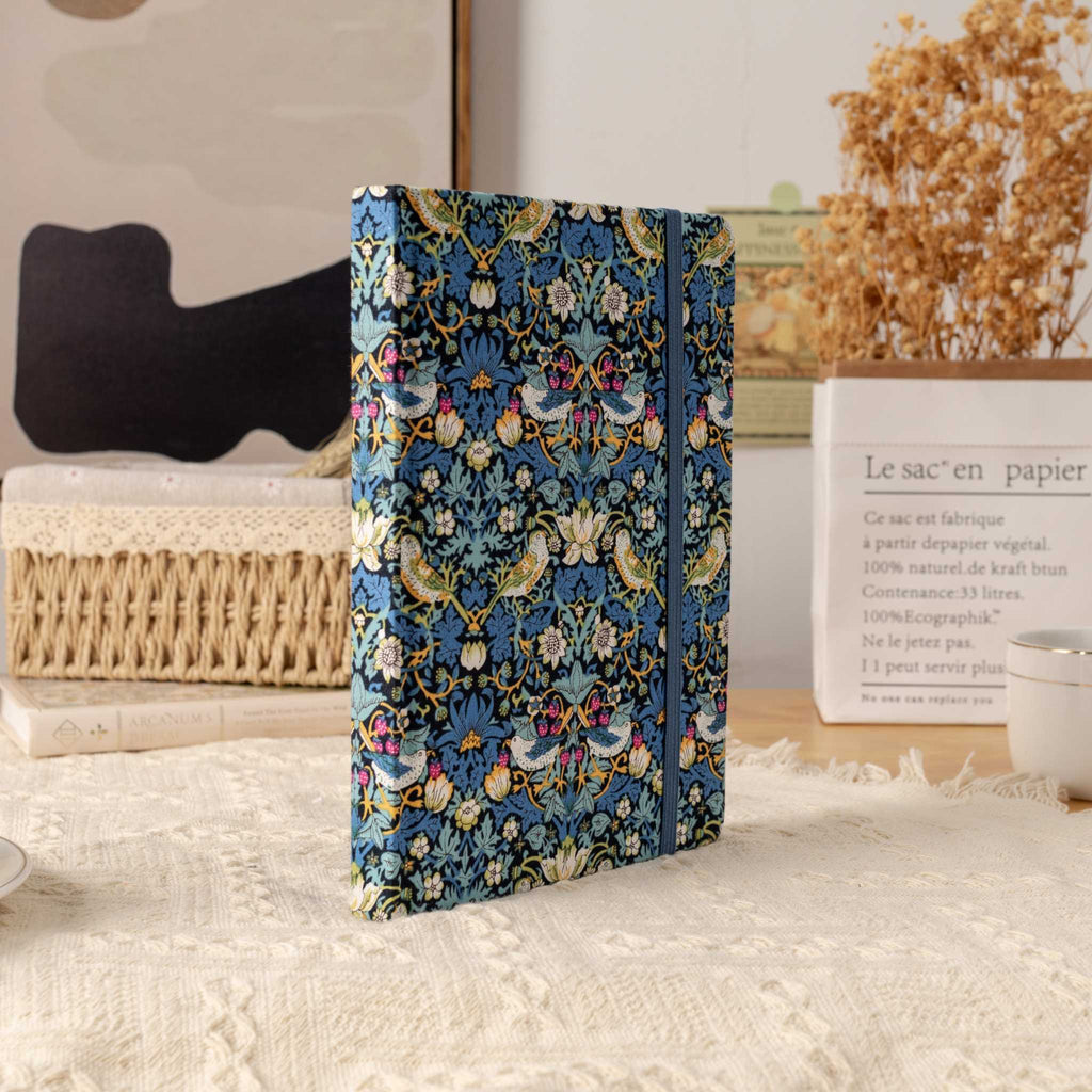 Blue bird pattern fabric notebook standing upright on a cream textured blanket, styled with a book and dried grass.
