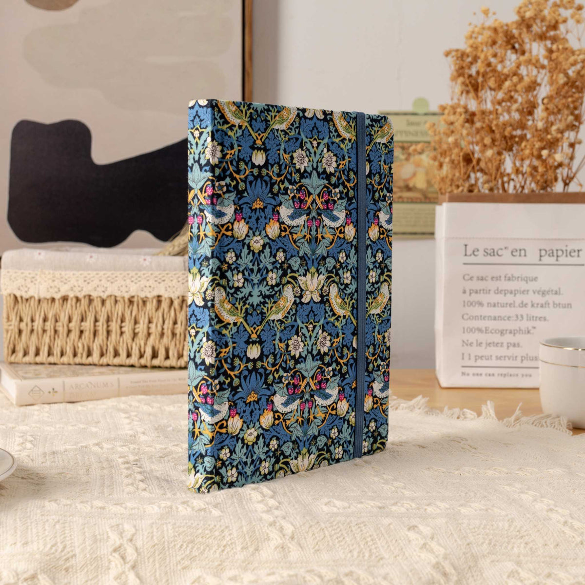 Blue bird pattern fabric notebook standing upright on a cream textured blanket, styled with a book and dried grass.
