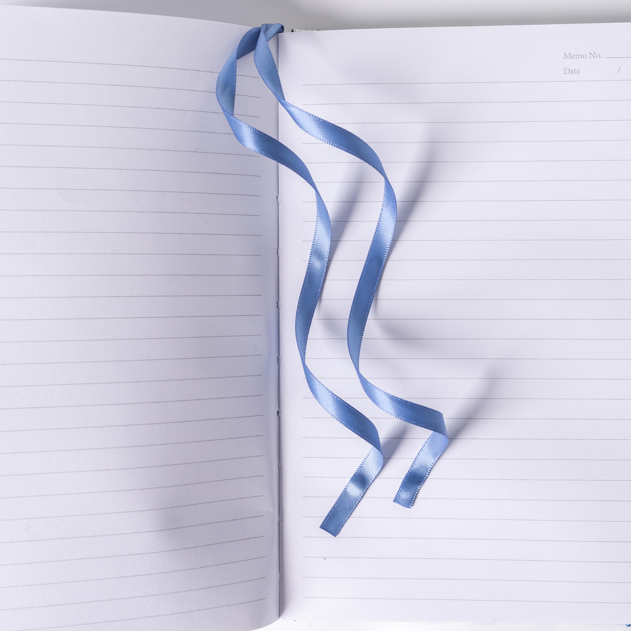Open A5 lined notebook showing clean horizontal pages with a light blue satin ribbon bookmark elegantly draped, and Memo No. and Date headers at the top right corner.