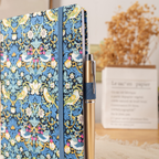 Side view of the blue bird fabric notebook standing upright, showing a blue and gold pen tucked into the side loop, styled with dried flowers and a book.