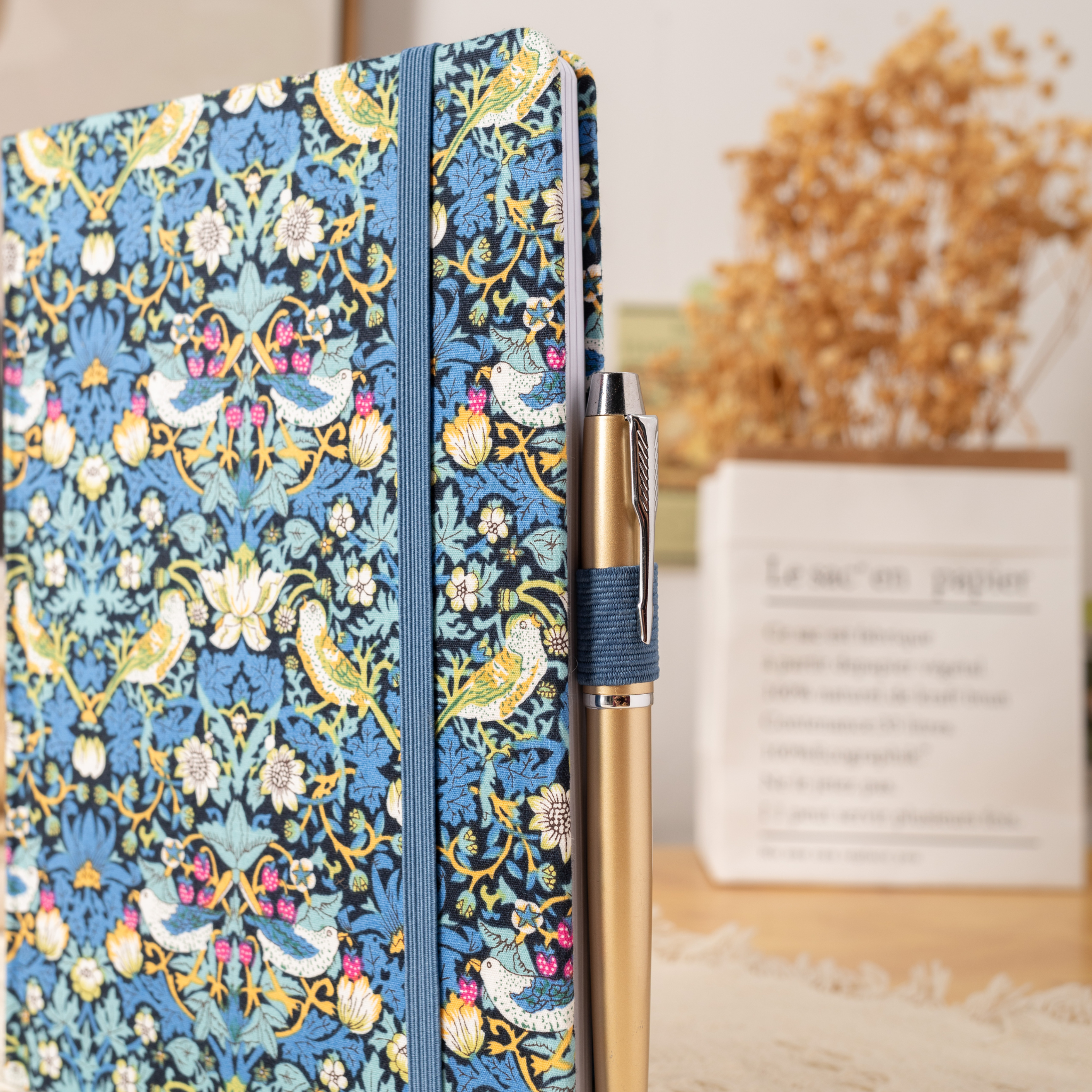 Side view of the blue bird fabric notebook standing upright, showing a blue and gold pen tucked into the side loop, styled with dried flowers and a book.